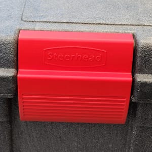 May include: A black plastic storage container featuring a prominent red latch mechanism. The red latch is embossed with the brand name Steerhead centered within a raised oval frame, and the lower portion of the latch has horizontal ribbed detailing.