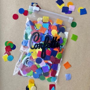 May include: A clear plastic bag filled with colorful confetti in various shapes, including circles and squares. The word "Confetti" is written in black script on the bag. Scattered confetti of different colors are on a brown surface.