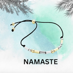 May include: A black string bracelet with gold and silver beads. The bracelet has a sliding knot closure. The word "NAMASTE" is written in black letters on a white background.