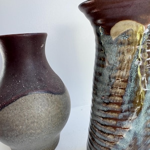 May include: Two ceramic vases with a rustic aesthetic. The vase on the left is a bulbous shape with a dark brown top and a speckled gray body. The taller vase on the right has a textured, ribbed design with brown, blue, and tan tones.