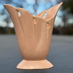 May include: A peach-colored ceramic vase with a flared, scalloped rim and a pedestal base. The vase has three circular openings near the top and vertical ridges. This decorative vase is suitable for displaying flowers or as a standalone accent piece.