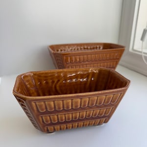 May include: Two brown ceramic rectangular planters with textured, repeating vertical and horizontal patterns. The planters have a glossy finish and are displayed on a white surface near a window.