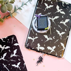 Könnte beinhalten: Eine schwarze Handyhülle mit einem Drachen- und Schwertmuster, ein silberner Handyhalter mit einem Zitat und ein Drachenanhänger. Der Handyhalter liest "A READER WITHOUT THEIR KINDLE IS A TRAGEDY".