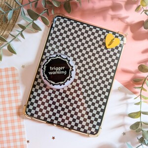 May include: A rectangular tablet case featuring a black and white checkered pattern. A circular pop-out grip is attached to the surface, displaying the text "trigger warning" inside a barbed wire border. A small yellow heart charm is also attached.