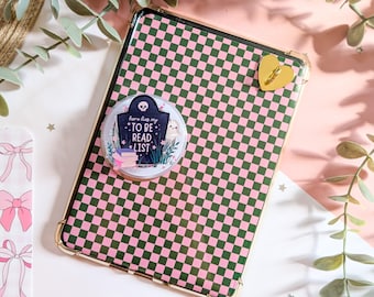 May include: A rectangular tablet case featuring a green and pink checkered pattern, adorned with a circular sticker depicting a tombstone with the text 'here lies my TO BE READ LIST', a stack of books, and a small ghost, plus a gold heart charm.