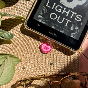 May include: A Kindle e-reader with a clear case displaying the book "Lights Out" and a pink heart-shaped charm that reads "Emotional Support Kindle." The scene includes eyeglasses, a straw mat, and greenery.