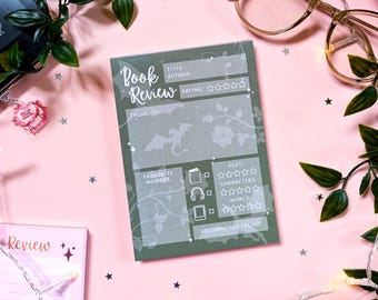 May include: A green book review notepad with the words "Book Review" at the top. The notepad has sections for title, author, rating, thoughts, favorite moments, plot, characters, world, and a recommendation. The background is pink with decorative elements.