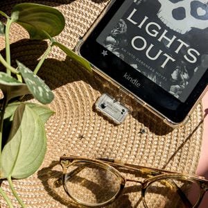 May include: A Kindle e-reader displaying the book cover for "Lights Out" with a skull graphic. The image also shows a pair of eyeglasses, a "Morally Grey Book Club" tag, and a plant on a woven straw mat.
