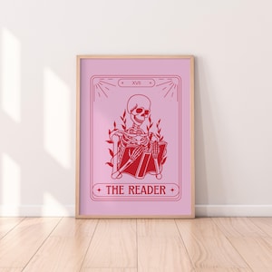 May include: A framed pink print with a red illustration of a skeleton reading a book. The print is labelled "THE READER" and includes the Roman numeral "XVII". The artwork is in a wooden frame.