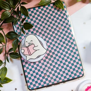 Peut inclure: Une coque de téléphone transparente avec un motif à carreaux roses et verts. Un support de téléphone en forme de fantôme blanc avec un dessin animé de fantôme lisant un livre rose est fixé à la coque. Des feuilles vertes et des confettis en forme d'étoiles sont en arrière-plan.