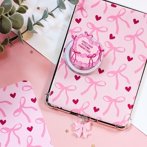 May include: Pink tablet case featuring a bow and heart design, a pop-up grip with the text "Cries Over Fictional Characters", and a bow charm. The case is on a pink surface with decorative elements.