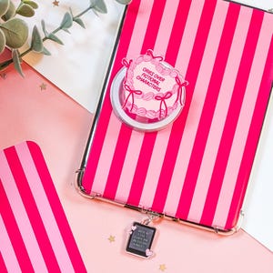May include: A pink and white striped phone case with a pop-up grip featuring a cake design that reads "Cries Over Fictional Characters." A charm with the text "When I die, bury me with my Kindle" hangs from the case.