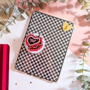 May include: A rectangular tablet case featuring a black and white checkered pattern with gold-toned corner accents. A circular sticker depicts a red heart-shaped cake with the text THRILLER LOVER and a small knife. A yellow heart charm is attached.