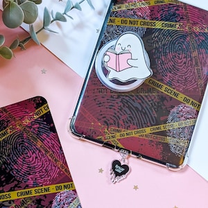Könnte beinhalten: Eine transparente Handyhülle mit einem roten und schwarzen Fingerabdruckmuster, gelbem "Crime Scene - Do Not Cross" Band und einem niedlichen Geist, der ein rosa Buch liest. Eine passende Karte und ein "Thriller Lover" Charm sind ebenfalls zu sehen.