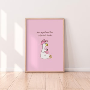 Könnte beinhalten: Ein gerahmter Druck mit einer Cartoon-Gans, die einen rosa Cowboyhut und eine Brille trägt und ein Buch hält. Die Gans ist vor einem rosa Hintergrund mit dem Text "just a girl and her silly little books" platziert. Der Rahmen hat eine helle Holzfarbe.
