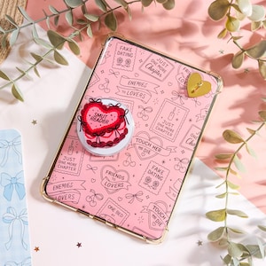 May include: A pink tablet case featuring a repeating pattern of book-themed phrases like 'Fake Dating', 'Enemies to Lovers', and 'Just One More Chapter'. A heart-shaped phone grip with the text 'Smut Reader' is attached to the centre of the case.