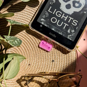 May include: A Kindle e-reader displaying the book cover "Lights Out" rests on a woven straw mat. A pink tag reading "Romantasy Book Club" is attached to the e-reader. Gold-framed glasses and green foliage are also visible in the image.