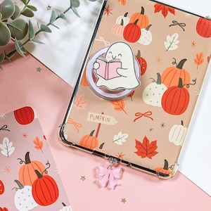 Könnte beinhalten: Eine transparente Handyhülle mit einem Herbstmuster aus Kürbissen und Blättern. Ein weißer, geisterförmiger Handyhalter liest ein rosa Buch. Ein rosa Schleifenanhänger hängt an der Hülle. Das Wort "PUMPKIN" steht auf einem Schild.