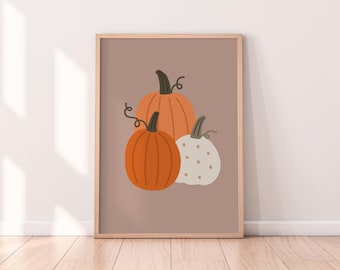 May include: A framed art print featuring three pumpkins in shades of orange and white against a muted brown background. The pumpkins have green stems and vines. The artwork is displayed on a wooden floor.