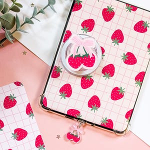 May include: A clear phone case with a pink strawberry pattern and a matching pop socket. The pop socket features a pair of strawberries with a pink bow. A strawberry charm hangs from the bottom of the case.