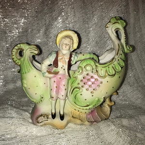 May include: A porcelain figurine of a boy in a green and pink outfit holding a paintbrush, sitting on a green and white ornate vase with gold accents.