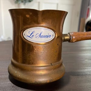 May include: A copper saucier pan with a cylindrical body, a flared rim, and a wooden handle. The front features an oval white enamel plaque with the blue text Le Saucier. The metal surface shows signs of oxidation and wear consistent with age.