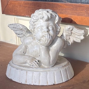 May include: A distressed white cherub statue with detailed wings, resting its chin on its hand. The statue has a round base and is made of a material that appears to be plaster or concrete. The cherub's expression is thoughtful.
