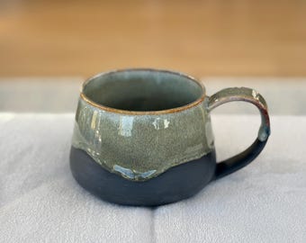 May include: A handmade ceramic mug with a dark blue base and a green upper section. The mug has a rounded shape with a handle and a brown rim. The mug is sitting on a white surface.