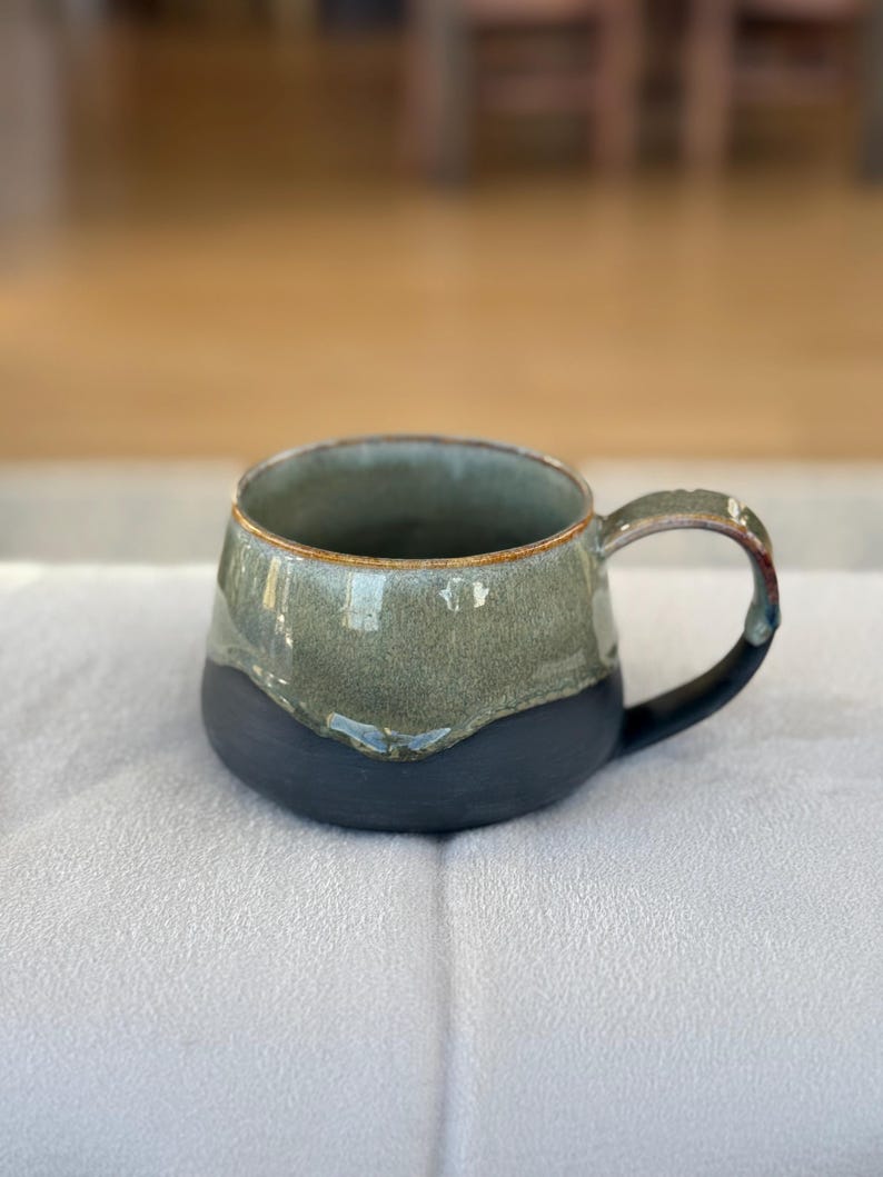 Op de afbeelding: Een handgemaakte keramische mok met een donkerblauwe basis en een groen bovenstuk. De mok heeft een ronde vorm met een handvat en een bruine rand. De mok staat op een wit oppervlak.