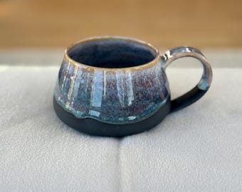 Può includere: Una tazza in ceramica fatta a mano con una glassa blu scuro e marrone. La tazza ha una base e un manico neri e un bordo dorato. La tazza è alta circa 10 cm e larga 13 cm.