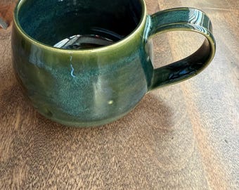 May include: A handmade ceramic mug with a glossy, emerald green glaze. The mug has a rounded body, a large handle, and a dark interior. The mug is sitting on a wooden surface.