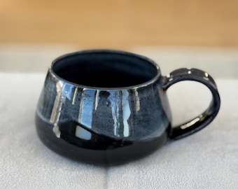 Op de afbeelding: Een handgemaakte keramische mok met een afgeronde body en een zwart handvat. De mok heeft een glanzende zwarte basis die overgaat in een donkerblauw-grijs met witte druppels. De binnenkant is diepblauw.
