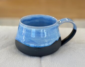 Op de afbeelding: Een handgemaakte keramische mok met een blauw geglazuurde bovenkant en een zwarte basis. De binnenkant is blauw en het handvat is zwart. De mok staat op een wit oppervlak.