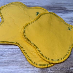 May include: Two yellow, petal-shaped cloth pads with dark green stitching around the edges. The pad on the top right has the letter "L" embroidered in green, and the pad on the top left has the letter "R" embroidered in green.