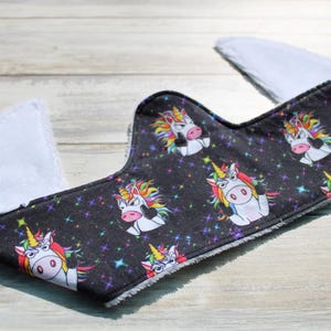 May include: A black bib with a pattern of white unicorns with rainbow manes and horns, set against a starry background. The bib has white, absorbent fabric on the edges and back, designed to catch spills and drool.