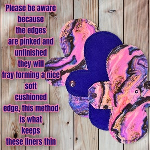 May include: Three reusable cloth liners featuring a pink, purple, and black marbled pattern with gold accents. The liners have dark purple fabric on the reverse side and include a snap closure. Text on the image explains the pinked, unfinished edges.