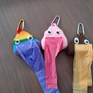 Op de afbeelding: Drie gehaakte poep-emoji-vormige hondenpoepzakhouders. De eerste is regenboogkleurig, de tweede is roze en de derde is bruin. Elke houder heeft glimmende ogen en een plastic zakdispenser.