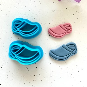 May include: Three blue and pink plastic cookie cutters shaped like shoes. The cutters are on a white surface with black speckles.