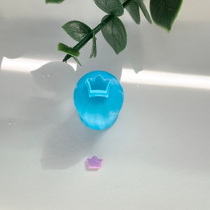May include: A light blue resin crown charm with a smaller purple resin crown charm on a white surface.