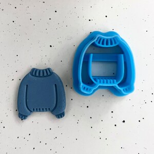 Könnte beinhalten: Zwei blaue Plastik-Ausstecher in Form von Pullovern. Der größere Ausstecher hat ein detailliertes Design mit einer gerippten Textur und einem umgeklappten Kragen. Der kleinere Ausstecher ist eine durchgehende Form mit einem einfachen Design.