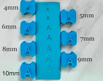 May include: A selection of blue letter "A" stamps in sizes from 4mm to 10mm, with a rectangular stamp also showing the letter "A". The sizes are labelled in millimetres. The background includes a marble surface and floral decorations.