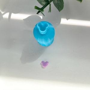 May include: Two small plastic figurines in the shape of Mickey Mouse. The larger figurine is blue and the smaller figurine is purple.