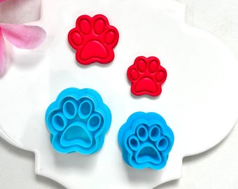 May include: Four paw print cookie cutters, two red and two blue, on a white ceramic tile background.