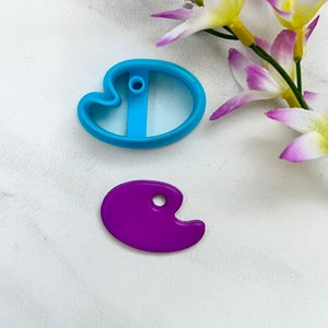 Puede incluir: Un cortador de galletas de plástico azul con forma de paleta de artista y un cortador de galletas de plástico morado con forma de paleta.