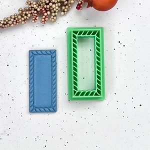 Puede incluir: Dos cortadores de galletas de plástico, uno azul y otro verde, con un diseño de cuerda alrededor de los bordes. Ambos cortadores son rectangulares.