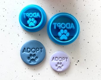 May include: Four blue and purple cookie cutters and fondant impressions with the word "ADOPT" and a paw print.