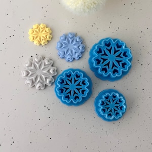 Puede incluir: Conjunto de seis cortadores de galletas con forma de copo de nieve de diferentes tamaños. Los cortadores están hechos de plástico azul y blanco. El cortador más pequeño es amarillo.