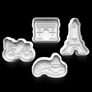 May include: Four white plastic cookie cutters in the shapes of a bicycle with bread, the Arc de Triomphe, the Eiffel Tower, and a croissant with coffee. The cutters are on a black background.
