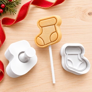 May include: A white plastic cookie cutter set for a stocking shape, including a plunger tool and a separate cutter frame, alongside a light brown stocking-shaped cookie on a white stick, arranged with a red ribbon and pine needles.