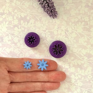 May include: Two purple flower-shaped cookie cutters and two small light blue flower-shaped objects. The cutters are round with a flower design. The small flowers are held on a finger. A sprig of purple flowers is in the background.